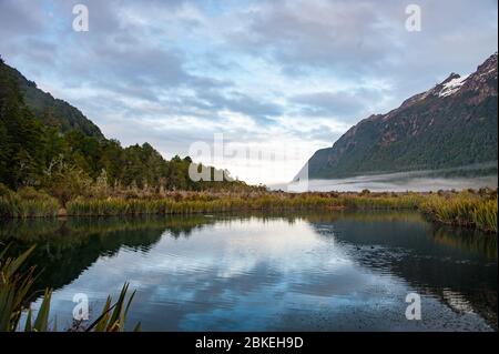 Sunrise, Mirror Lakes in Fiordland National Park, New Zealand. Mist hangs low in the valley with mountain and  colourful sky reflections on the water. Stock Photo