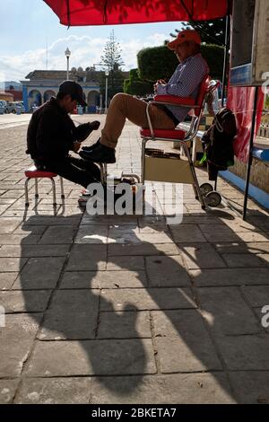 Young shoe shine boy cleaning shoes for a man in the Plaza de Teopisca at sunset. Stock Photo