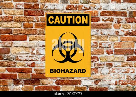 Old yellow warning sign screwed to a brick wall. In the middle of the panel, there is a biohazard symbol and the message is saying 'Caution, Biohazard Stock Photo