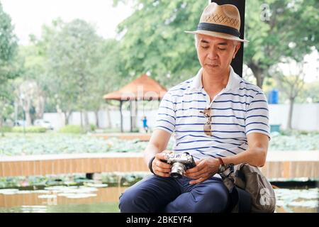 Elderly Chinese man resting by pond in park and checking photos he made on new digital camera during walk Stock Photo
