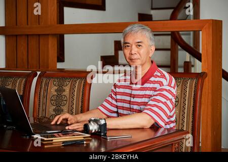 Mature Chinese photographer working on laptop at home and editing photos on laptop Stock Photo