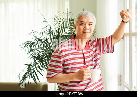 Happy mature Asian man standing at window and drinking morning coffee at home Stock Photo