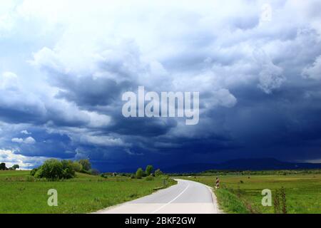 Road in the field, stormy clouds over the hill in background Stock Photo