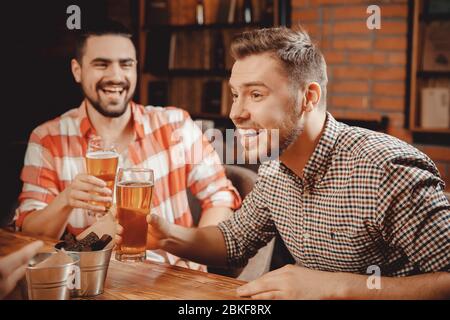 Friends young bearded men have fun in bar, clanging glasses of beer, laughing, on table they snacks cans that mimic buckets. Stock Photo