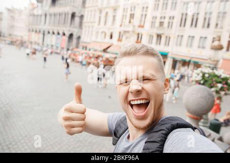 Young bearded blond guy smile traveler to Europe with backpack taking selfie on Grand place and showing Thumbs up thumb. Stock Photo