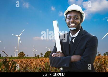 African engineer wearing white hard hat standing with against wind turbine on sunny day Stock Photo