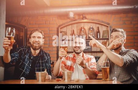 Three friends sitting in bar, drinking beer, watching match on TV, rooting for ours. Guy crossed his arms stretching out index fingers Stock Photo