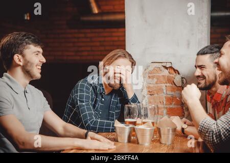 Bearded guys plaid shirts laughing at table in Irish pub drinking beer, young man with long blond hair facepalm, peeling brick wall background Stock Photo