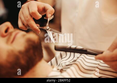 Barber hands trimmed beard of man, focus on scissors closeup. Background is blurred Stock Photo