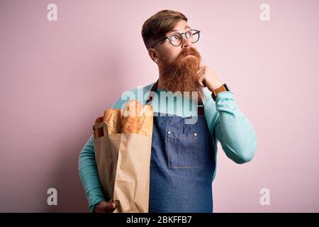 Irish redhead baker man with beard holding groceries paper bag of bread over yellow background serious face thinking about question, very confused ide Stock Photo