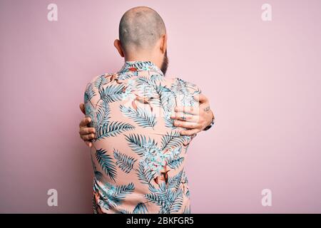 Handsome bald man with beard and tattoo wearing casual floral shirt over pink background Hugging oneself happy and positive from backwards. Self love Stock Photo