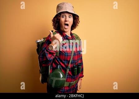 Middle age curly hair hiker woman hiking wearing backpack and water canteen using binoculars Surprised pointing with hand finger to the side, open mou Stock Photo