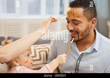 Young woman and little girl having fun painting colourful gouache strokes on face of happy young man Stock Photo