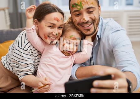 Young husband and wife taking funny selfie on smartphone with their little daughter with gouache on their faces Stock Photo