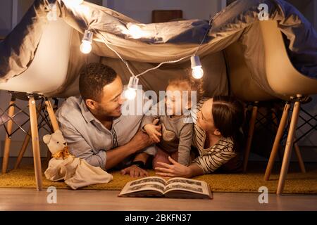 Young parents having fun with their little daughter in handmade tent at home in evening, horizontal shot Stock Photo
