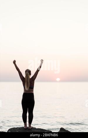 Young woman standing by the sea with arms raised in triumph Stock Photo