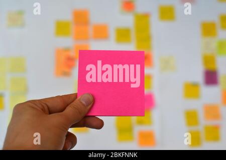 A hand is holding a pink empty sticker and there is a Kanban board of agile methodology on the background, which is a developing trend in IT Stock Photo