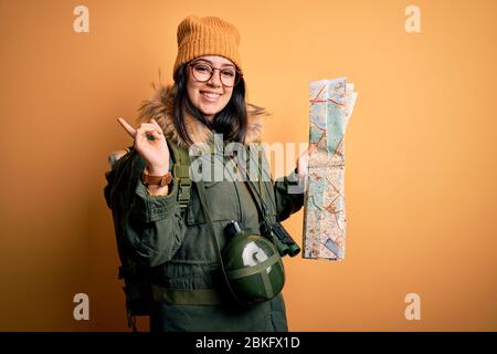 Young hiker woman wearing hiking backpack looking at tourist map over yellow background very happy pointing with hand and finger to the side Stock Photo