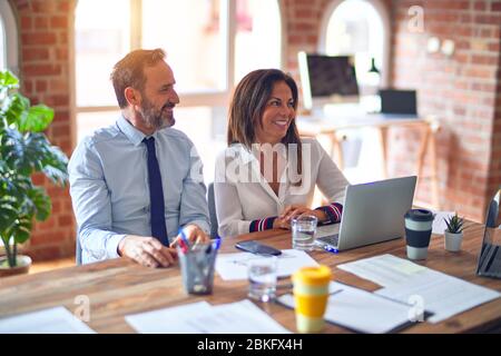 Middle age beautiful business workers working together using laptop at the office looking away to side with smile on face, natural expression. Laughin Stock Photo