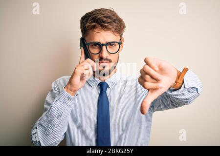 Young businessman having a conversation talking by smartphone over white background with angry face, negative sign showing dislike with thumbs down, r Stock Photo