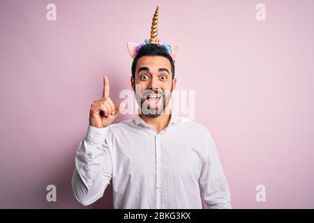 Young handsome man with beard wearing funny unicorn diadem over pink background pointing finger up with successful idea. Exited and happy. Number one. Stock Photo