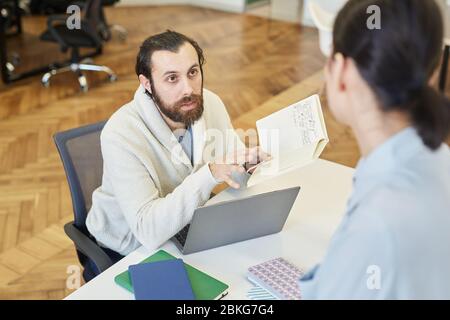 Young man sitting at office desk sharing his ideas to unrecognizable colleague, horizontal high angle shot Stock Photo