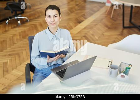 Horizontal high angle portrait of Caucasian woman wearing light blue shirt and jeans sitting at office desk holding notebook, looking at camera Stock Photo