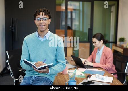 Young African American man with short dreadlocks sitting relaxed on office table holding notebook and pen, looking at camera smiling Stock Photo