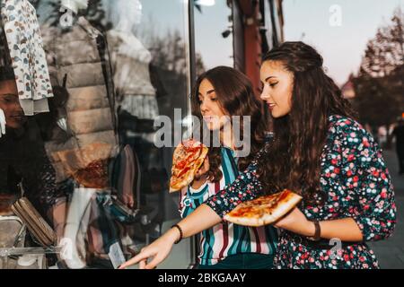 Two beautiful girl talking and eating pizza outdoors, and pointing finger in the shop window. Consumerism, shopping, lifestyle concept Stock Photo