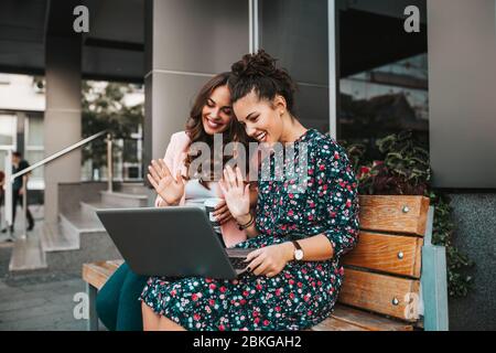 Two beautiful young women friends waving hand and talking while having a video call with their friends on a laptop in a city street. Stock Photo