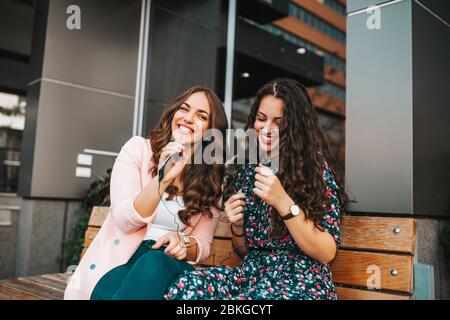 Two pretty happy cheerful  women - friends using mobile phone singing and listening music together with earphones. Stock Photo