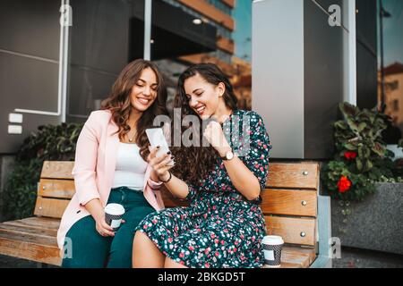 Two happy women friends celebrating success while reading good news on a smartphone outdoors in the city street.