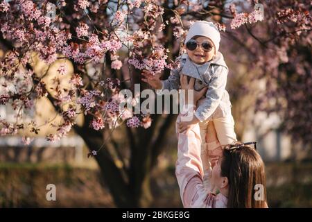 Beautiful mom with adorable daughter stand by pink blossoming tree. Happy family spend time in Mother's Day. Cute little girl laughing Stock Photo