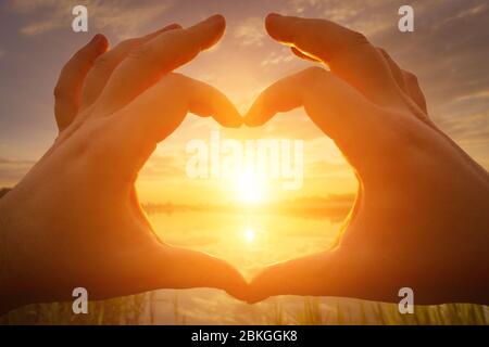 Hands in the shape of heart against the sunset or sunrise over the lake or river in the summer with a cloudy sky background. Landscape. Stock Photo