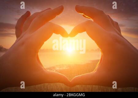 Hands in the shape of heart against the sunset or sunrise over the lake or river in the summer with a cloudy sky background. Landscape. Stock Photo