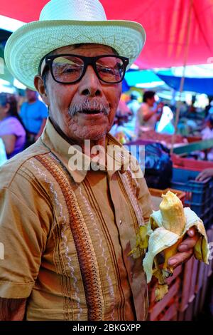 Portrait of an old man with a straw hat eating a banana in the Zaachila market. Stock Photo