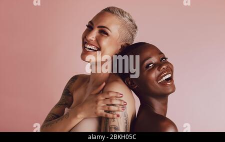 Two women with buzz cut hairstyle standing back to back and smiling at camera. Female models with beautiful skin smiling together over beige backgroun Stock Photo