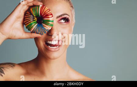 Close-up of a woman with short hair holding donut in front of her eye and sticking out her tongue. Beautiful female with colorful donut on grey backgr Stock Photo