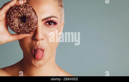 Close-up of a woman with short hair looking through a donut and sticking out her tongue. Beautiful female with a donut on grey background. Stock Photo