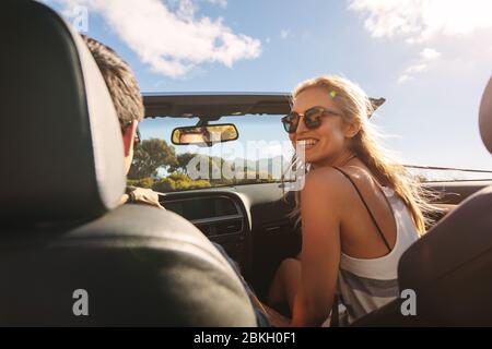 Rear view of couple sitting inside a convertible car. Woman looking at her boyfriend driving the car. Stock Photo