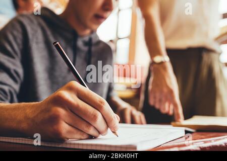 Close up of teenage boy writing notes sitting at desk during lecture with female teacher pointing at a book. Focus on hand writing in book in classroo Stock Photo