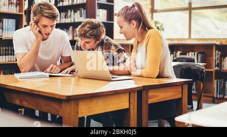 Students working on school project in library, sitting at table with laptop and reading at book. Young university students studying at library. Stock Photo