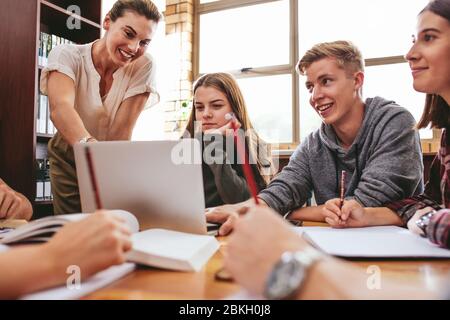 Female teacher working with college students in library. Group of students studying in school library with a teacher helping them. Stock Photo