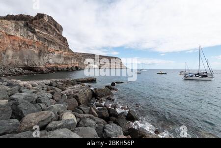 Yachts moored off the rocky southern coastline of Gran Canaria taking refuge in the shelter of the cliffs in the bay near Puerto de Mogan. Stock Photo