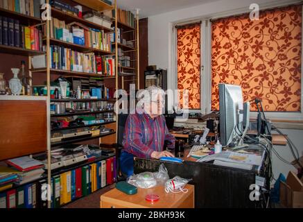 Elderly white haired man working at home on the desktop computer in a mess. Bookshelf with many books and antiviral masks ready Stock Photo