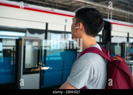 Close up of young man listening to music with wireless earpods while commuting by train at station. Asian guy enjoying music on the go. Stock Photo