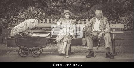 Panoramic, monochrome view of senior 1940s couple - grandparents sitting on bench, baby in vintage pram, Severn Valley Railway heritage station. Stock Photo