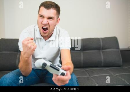 Young man sit in room during quarantine. Emotional guy playing computer games. Male person hold joystick in hand and cheer with pleasure. Screaming Stock Photo