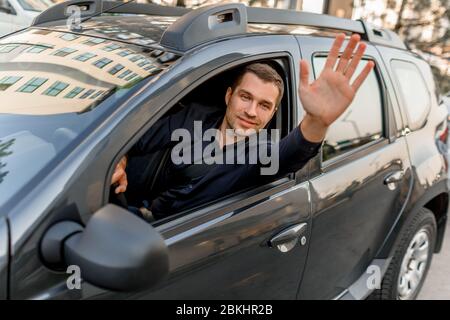 A young man in a shirt sits in his car and waves to someone. Taxi driver looks outside and smiles. Urban environment and traffic Stock Photo