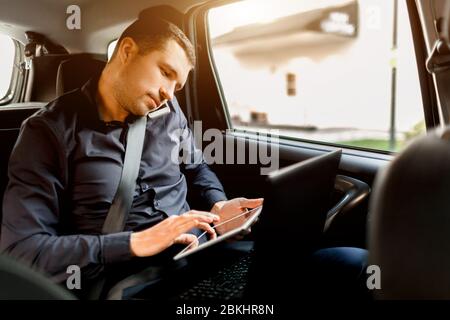 Busy businessman in a taxi. Multitasking concept. Passenger rides in the back seat and works simultaneously. Speaks on a smartphone and uses a laptop Stock Photo